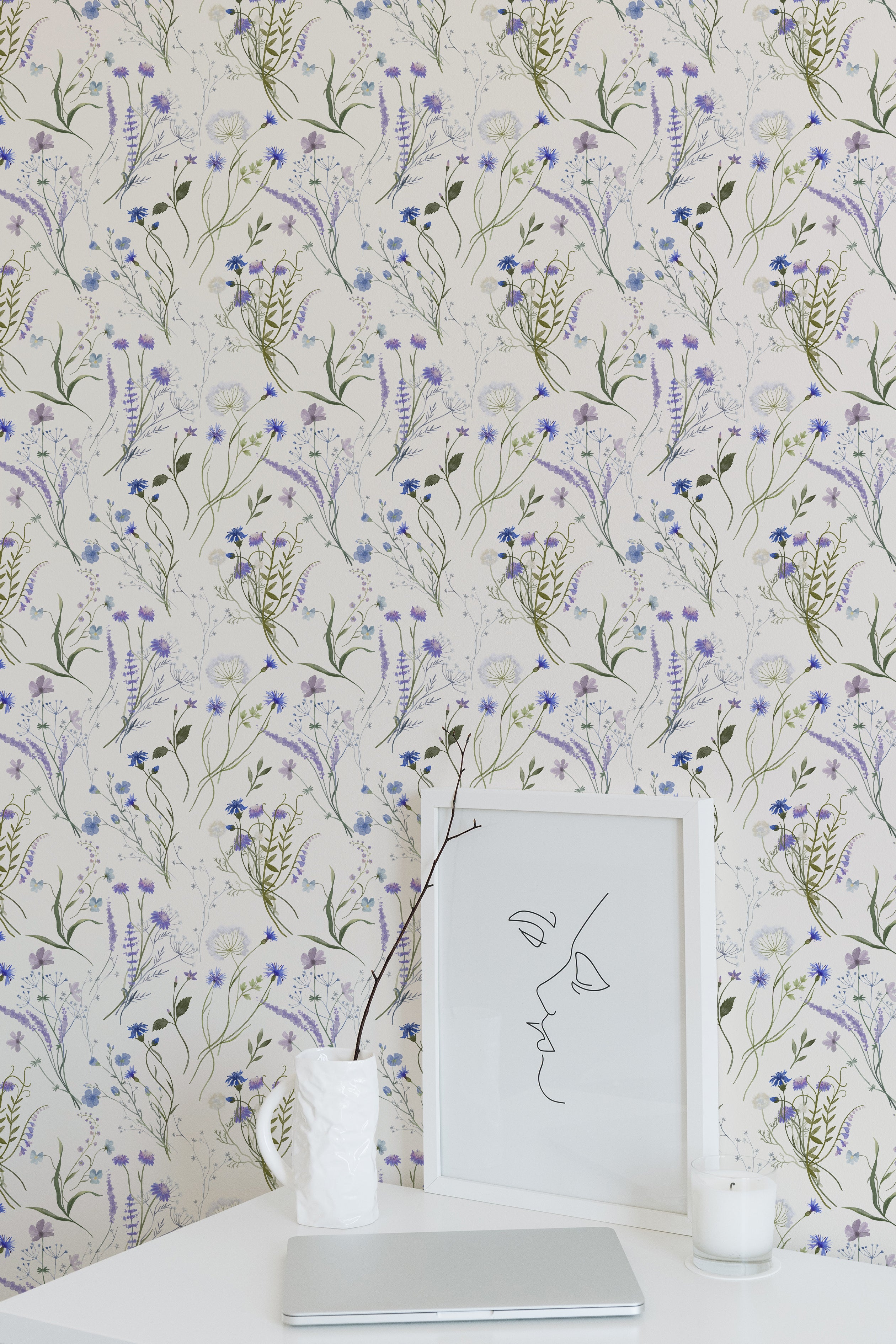 An elegant workspace showcasing the Wildflower Bouquet Wallpaper, featuring a beautiful array of hand-painted wildflowers in shades of blue and purple with green foliage. A minimalist desk with a framed line art and a decorative vase complements the floral backdrop.