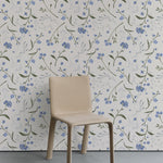 A simple modern chair stands against a wall adorned with Wildflower Bouquet Kids Wallpaper, featuring a delightful pattern of blue wildflowers and green foliage on a white background. The scene conveys a serene and natural setting, perfect for a peaceful interior.