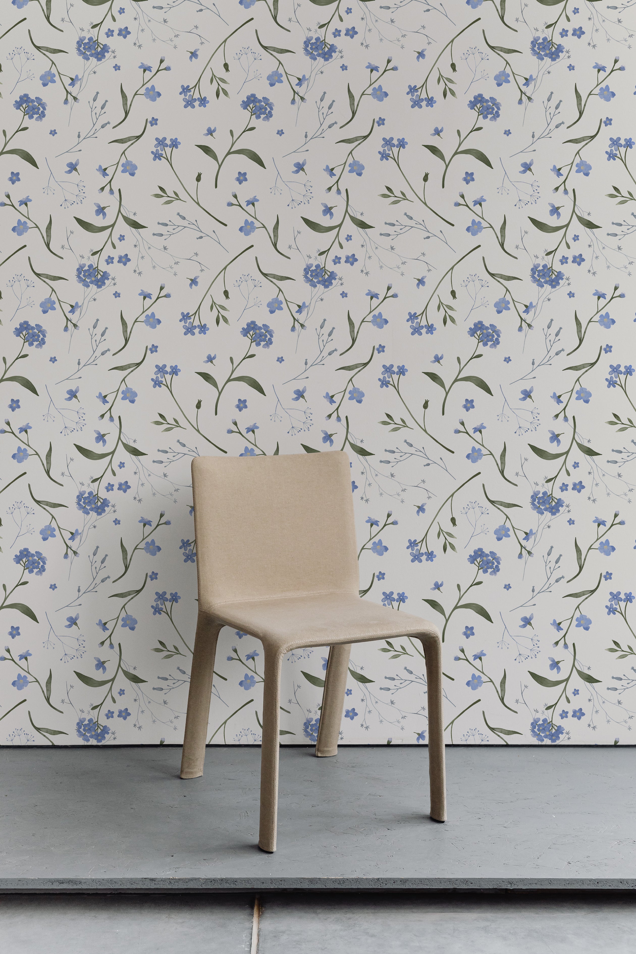 A simple modern chair stands against a wall adorned with Wildflower Bouquet Kids Wallpaper, featuring a delightful pattern of blue wildflowers and green foliage on a white background. The scene conveys a serene and natural setting, perfect for a peaceful interior.