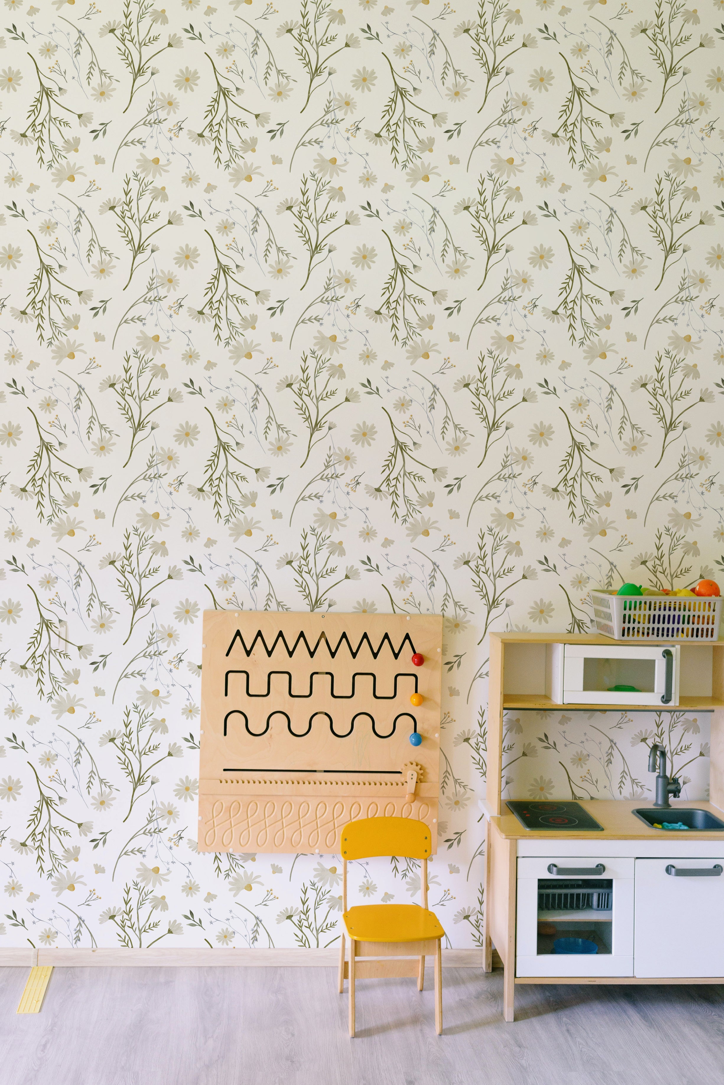 A cozy children's playroom with Daisy Bouquet Wallpaper featuring delicate daisies and foliage in soft greens and yellows on a white background. The room includes a child-sized wooden chair in yellow, a matching wooden play kitchen, and an educational wooden wall-mounted activity board with sliding beads and shapes