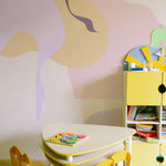 A vibrant and playful children's play area featuring the Contemporary Art - Abstract Wall Mural, with flowing abstract shapes in shades of yellow, purple, and pink. The space is furnished with a yellow child-sized table and chairs, and a yellow storage unit, creating a stimulating environment for creativity.