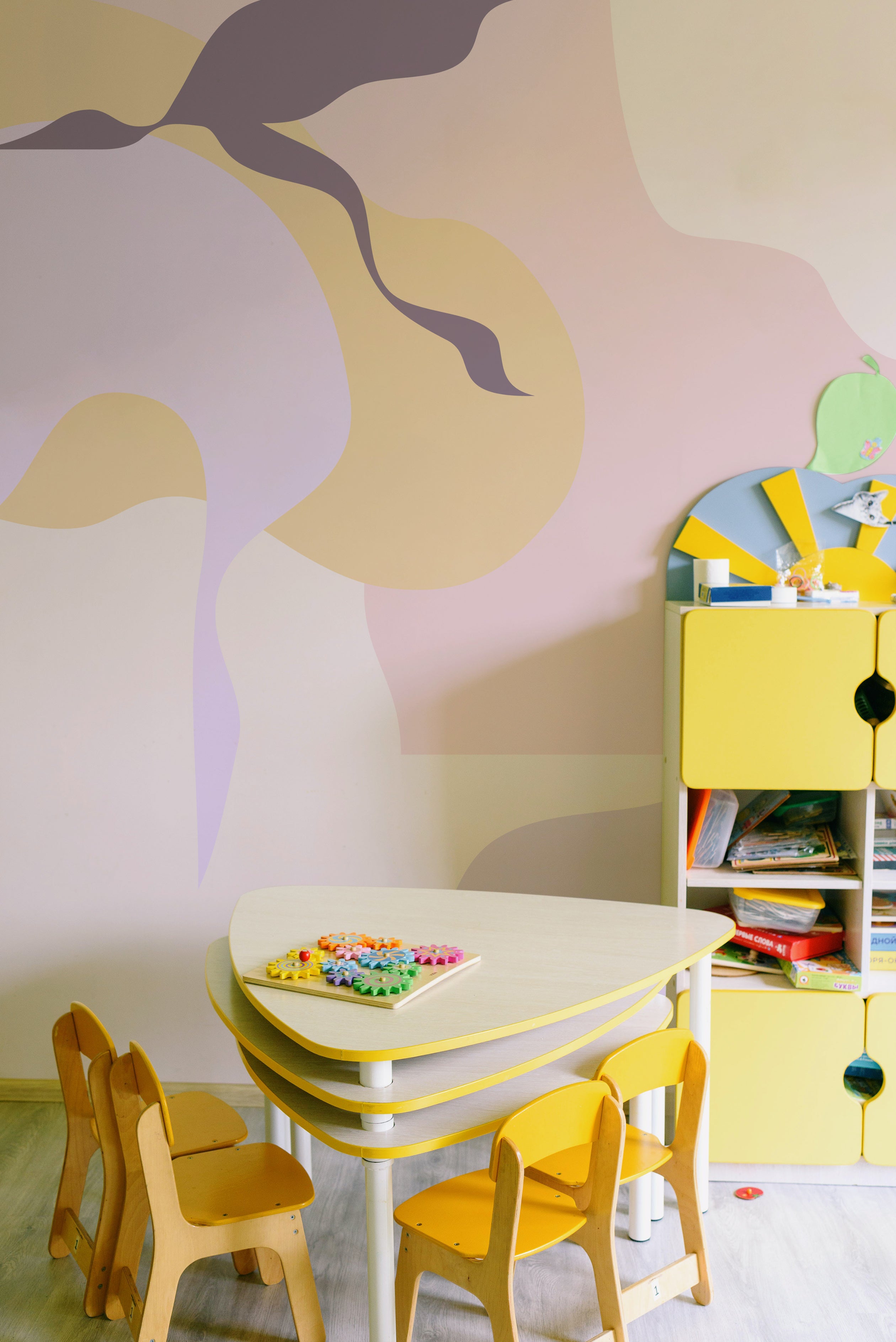 A vibrant and playful children's play area featuring the Contemporary Art - Abstract Wall Mural, with flowing abstract shapes in shades of yellow, purple, and pink. The space is furnished with a yellow child-sized table and chairs, and a yellow storage unit, creating a stimulating environment for creativity.