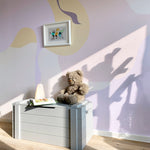 A sunny corner of a room with the Contemporary Art - Abstract Wall Mural providing a dynamic backdrop. The wall mural's organic abstract shapes in soft hues contrast beautifully with the sunlight casting shadows of tree branches, accompanied by a teddy bear sitting on a grey storage chest.