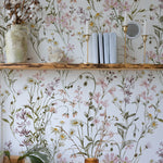 Interior scene with a section of wall covered in WildFloral Wallpaper displaying a delicate watercolor-style pattern of wildflowers in shades of pink, white, and yellow with green foliage. A rustic wooden shelf holds decorative items such as books, a vase with cotton branches, and brass candlesticks, blending with the floral design.