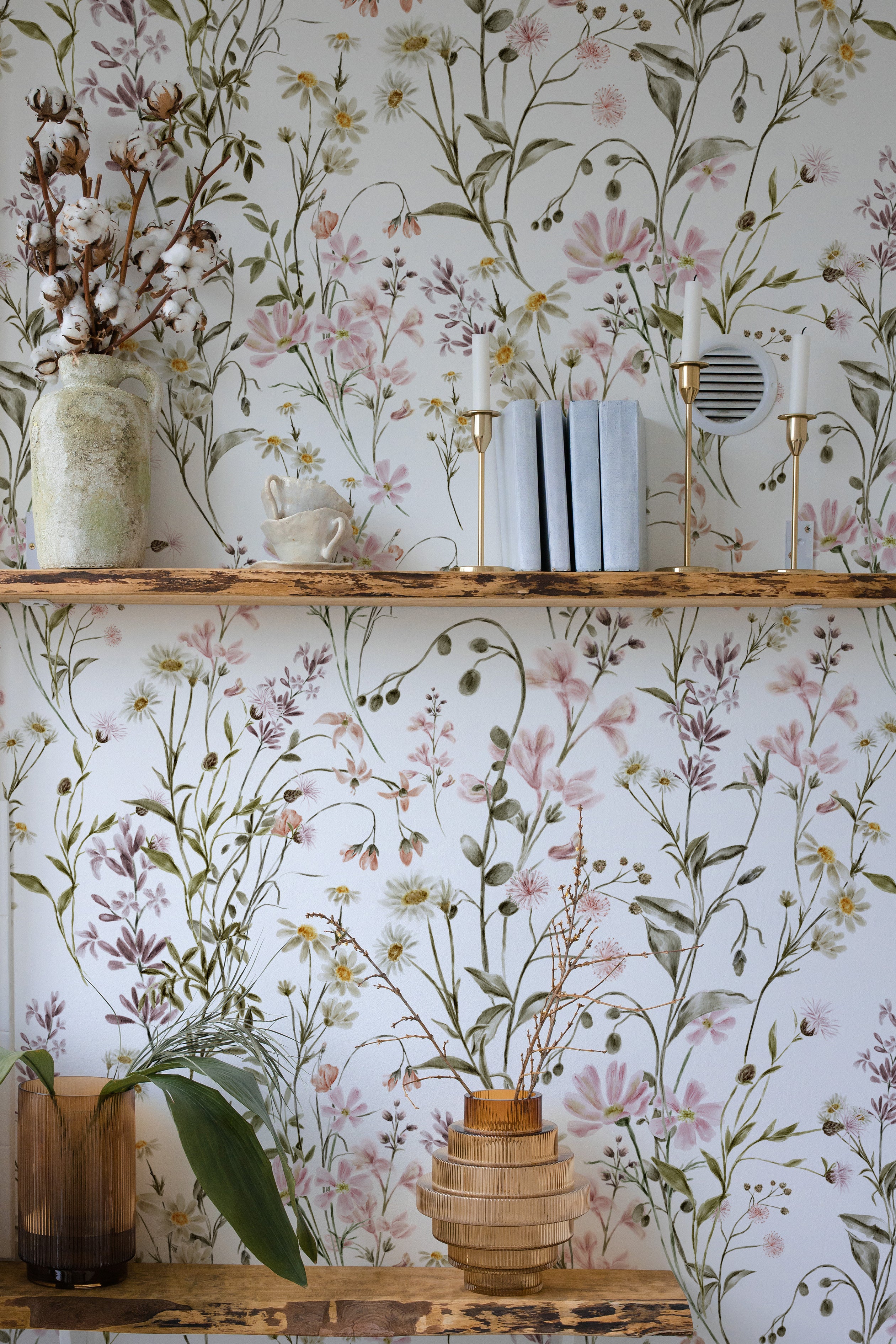 Interior scene with a section of wall covered in WildFloral Wallpaper displaying a delicate watercolor-style pattern of wildflowers in shades of pink, white, and yellow with green foliage. A rustic wooden shelf holds decorative items such as books, a vase with cotton branches, and brass candlesticks, blending with the floral design.