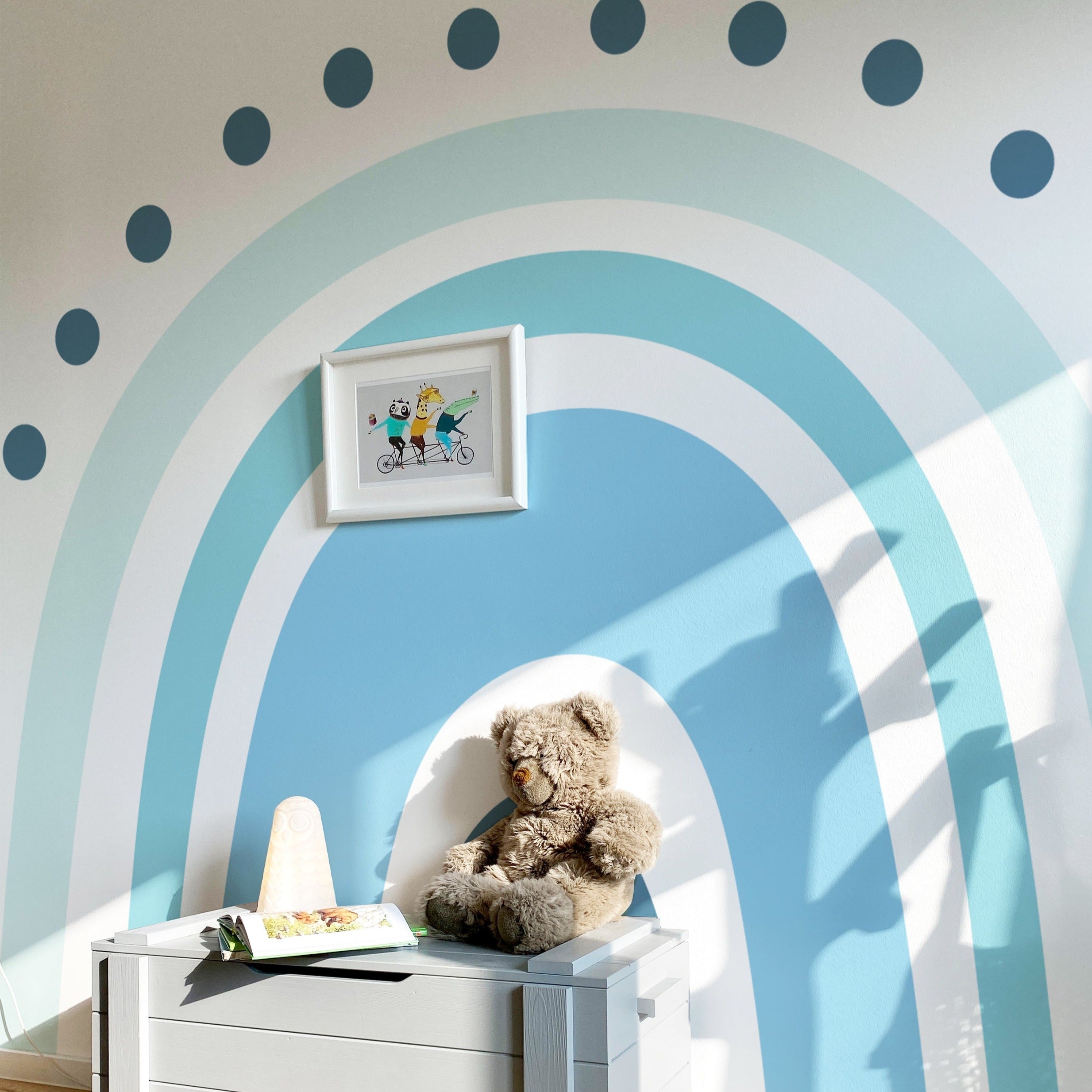 A child’s room adorned with the Rainbow Wallpaper Mural - Aurora, creating a joyful and playful atmosphere. The mural includes large blue and turquoise arches with a polka dot pattern, complemented by a framed illustration of cartoon characters on a bike, a teddy bear, and children's books, offering a vibrant and inviting space.