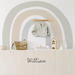 A stylish nursery room with a large Boho Rainbow Wallpaper Mural above a wooden wainscoting. The mural features soft pastel colors and is used as a backdrop for children’s accessories including a jacket, bag, and toys. Below the mural, the name "William" is stylized in metallic letters on the wooden panel.