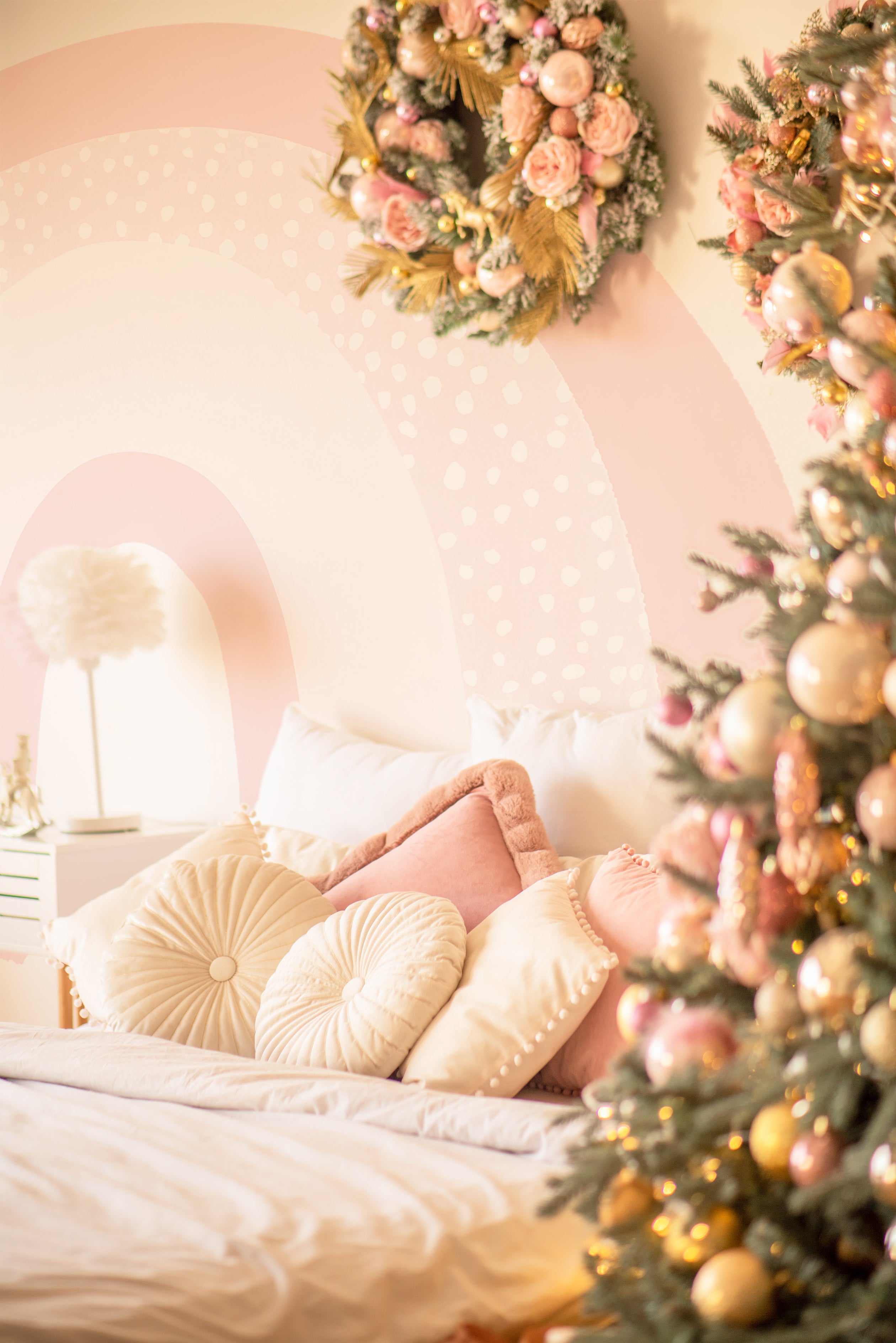 A festive holiday bedroom scene with Cute Rainbow Wallpaper Mural - Celeste adorning the wall. The mural features soft pastel rainbow arches with a dotted pattern, enhancing the warmth of the room decorated with Christmas elements, including a fully adorned tree and floral wreath.