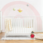 A nursery showcasing the Cute Rainbow Wallpaper Mural - Celeste, providing a playful and sweet backdrop. The wallpaper features soft pastel arches with a polka dot design, complemented by a white crib, a classic dresser, and whimsical hanging planets, creating a dreamy space for a child.