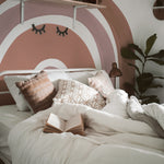 A cozy bedroom setting showcasing the Aesthetic Rainbow Wallpaper Mural - Luna. The mural features large, soft arches in muted tones of brown and pink, providing a warm backdrop. Decorated with plants and cozy bedding, this space reflects a tranquil and stylish ambiance.