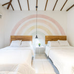 A serene bedroom with two single beds and wooden headboards against a wall featuring a large pastel rainbow mural with soft arcs of pink, peach, yellow, and green hues.
