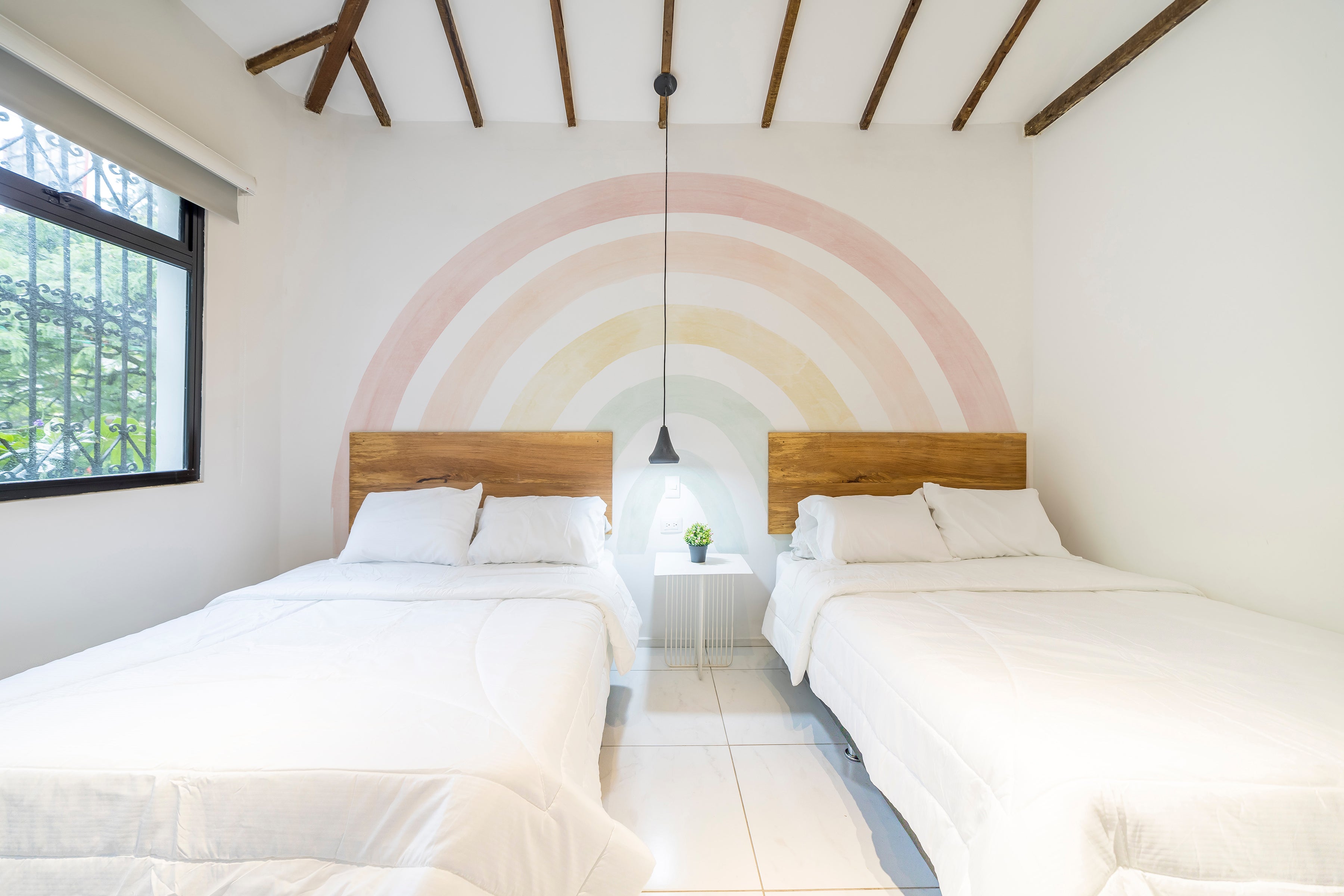 A serene bedroom with two single beds and wooden headboards against a wall featuring a large pastel rainbow mural with soft arcs of pink, peach, yellow, and green hues.