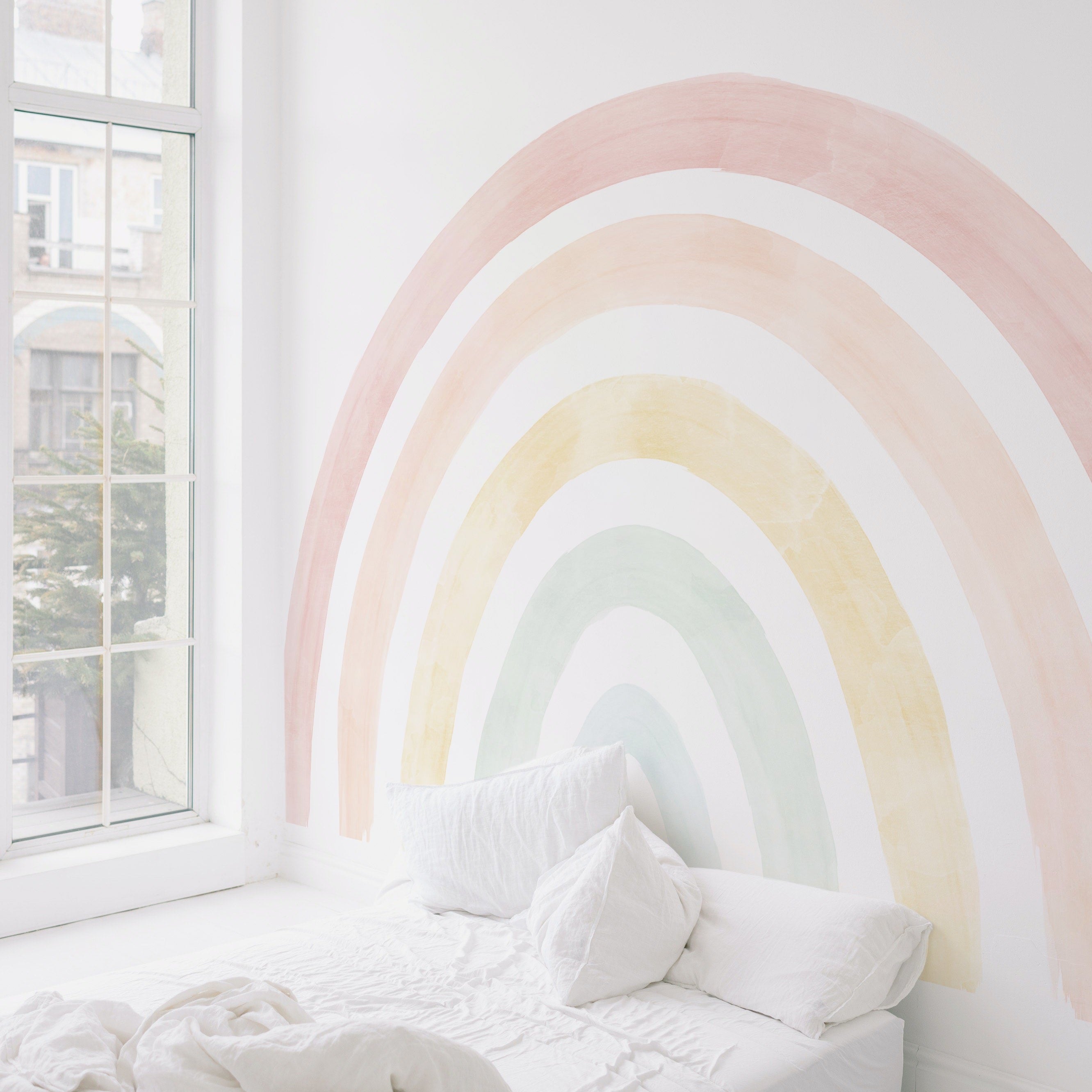 A cozy corner of a room lit by natural light through a large window, with the pastel rainbow mural providing a soothing backdrop to a simple white bed and linens.