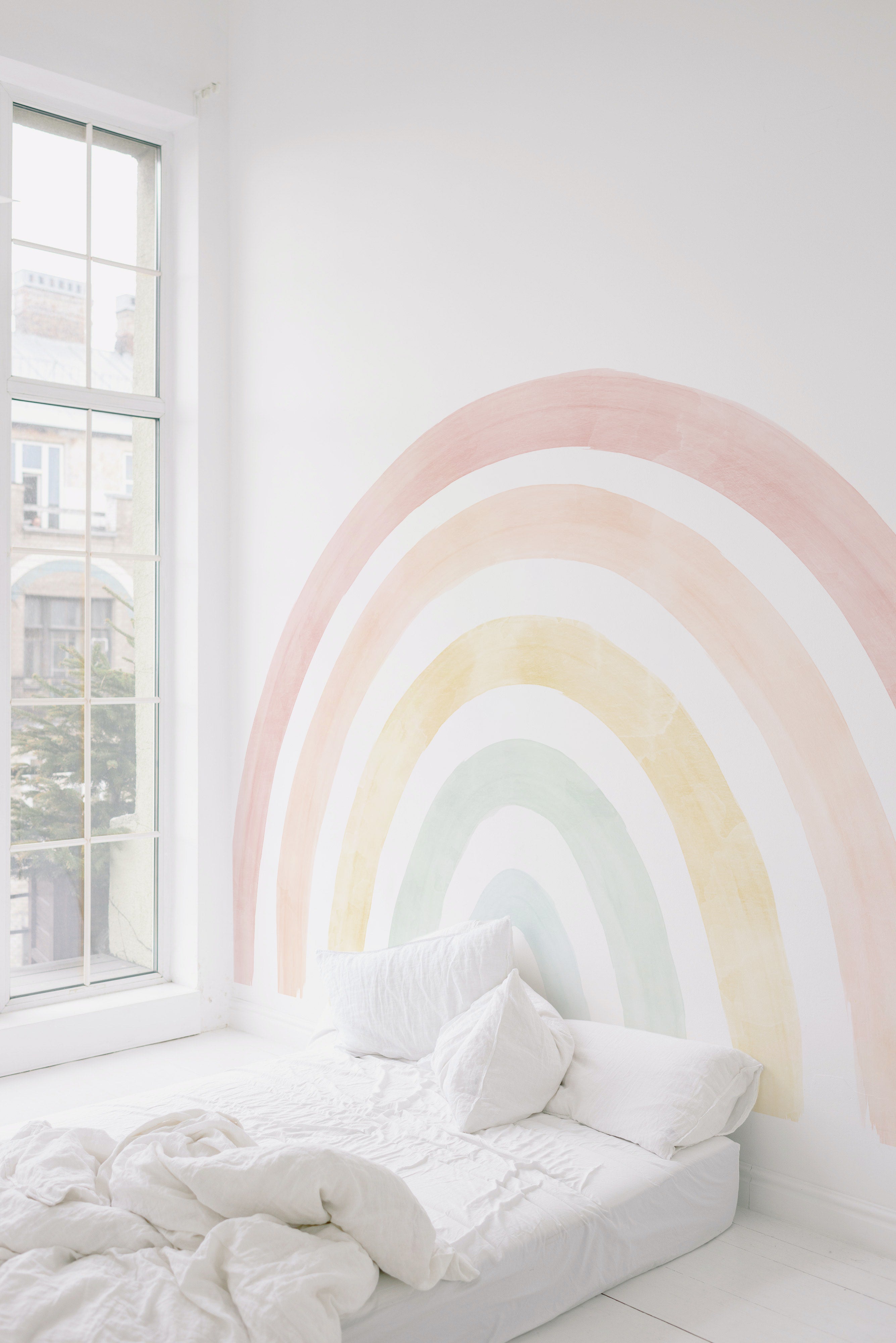 A cozy corner of a room lit by natural light through a large window, with the pastel rainbow mural providing a soothing backdrop to a simple white bed and linens.
