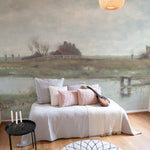 A cozy living room setting showcasing the River Landscape - Vintage Mural on a feature wall. The mural depicts a serene rural scene with thatched cottages by a river, under a vast, cloudy sky, offering a peaceful and picturesque view. Furnished with soft pillows on a stylish daybed, the room exudes a tranquil, welcoming atmosphere.