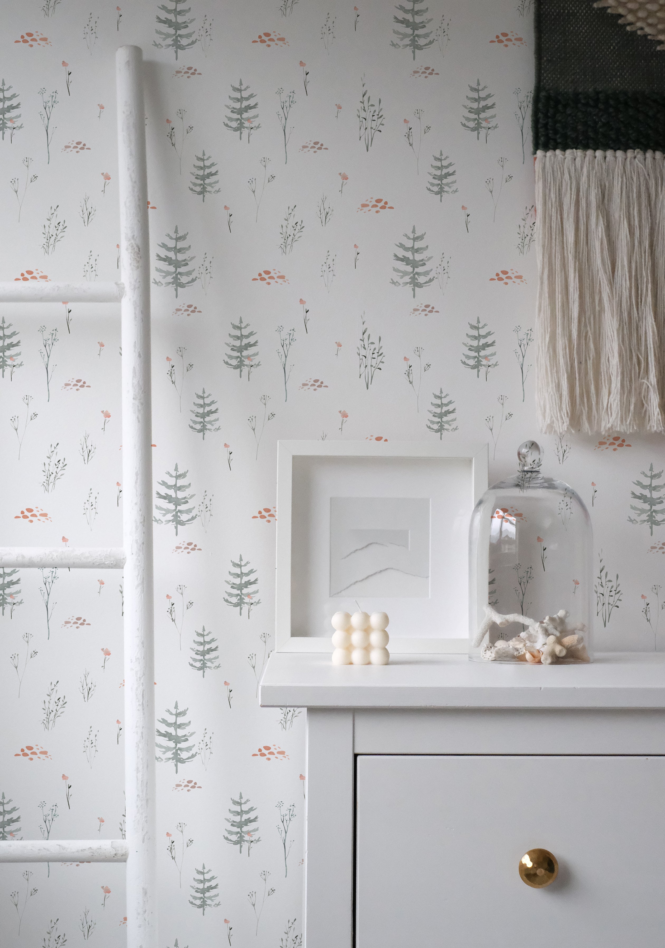 A styled corner of a room showcasing the Pine Tree Wallpaper. The wall is adorned with soft gray pine trees and small foliage, complemented by a white fireplace mantel displaying a minimalist art frame and a decorative glass cloche containing sea shells.