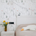 a bedroom corner with "Pine and Birch Wallpaper" on the wall. The light and airy wallpaper, depicting delicate birch and pine trees alongside gentle floral accents, brings a sense of outdoor tranquility to the interior space. A vase with bright yellow flowers on a bedside table adds a pop of color to the serene setting.