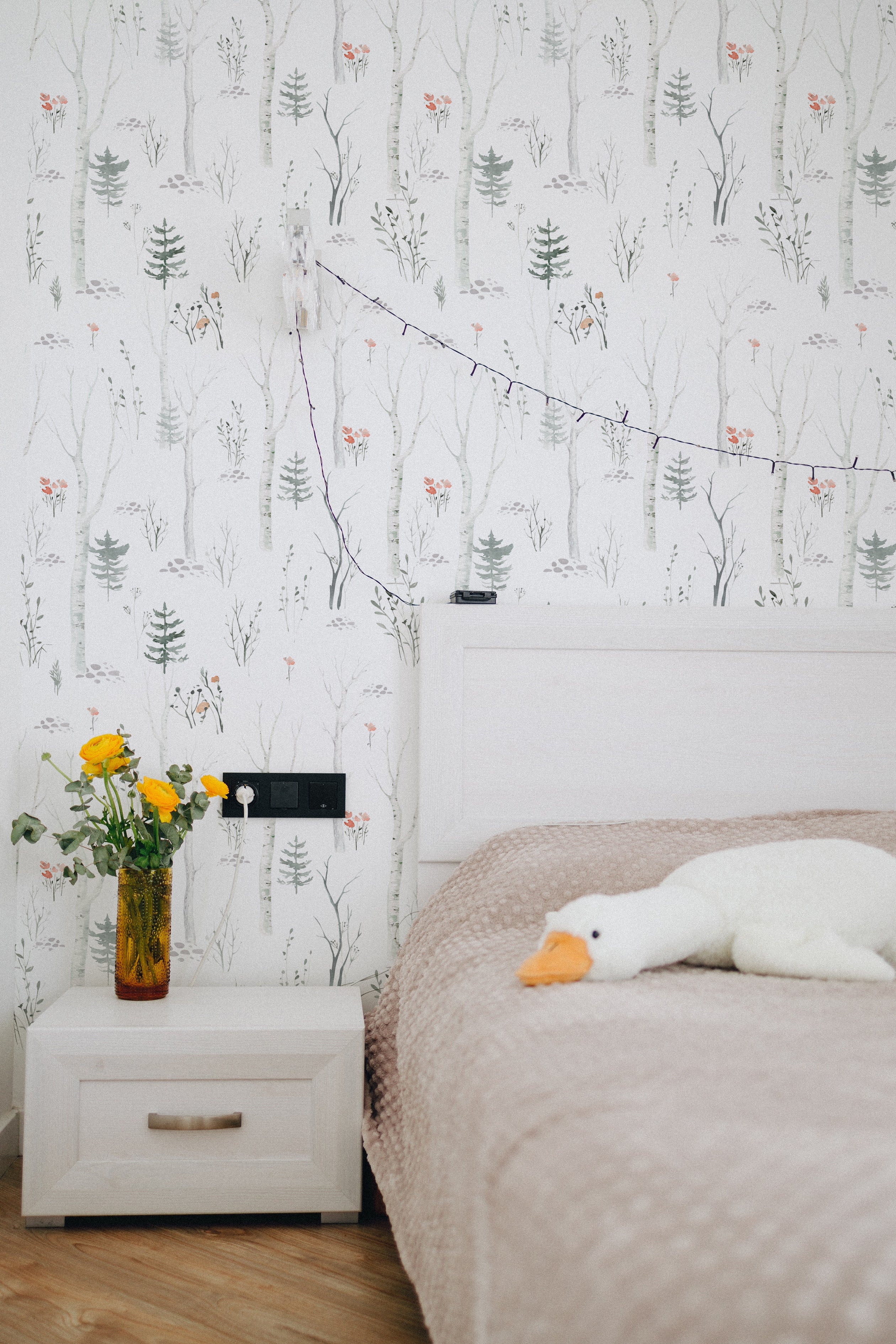 a bedroom corner with "Pine and Birch Wallpaper" on the wall. The light and airy wallpaper, depicting delicate birch and pine trees alongside gentle floral accents, brings a sense of outdoor tranquility to the interior space. A vase with bright yellow flowers on a bedside table adds a pop of color to the serene setting.