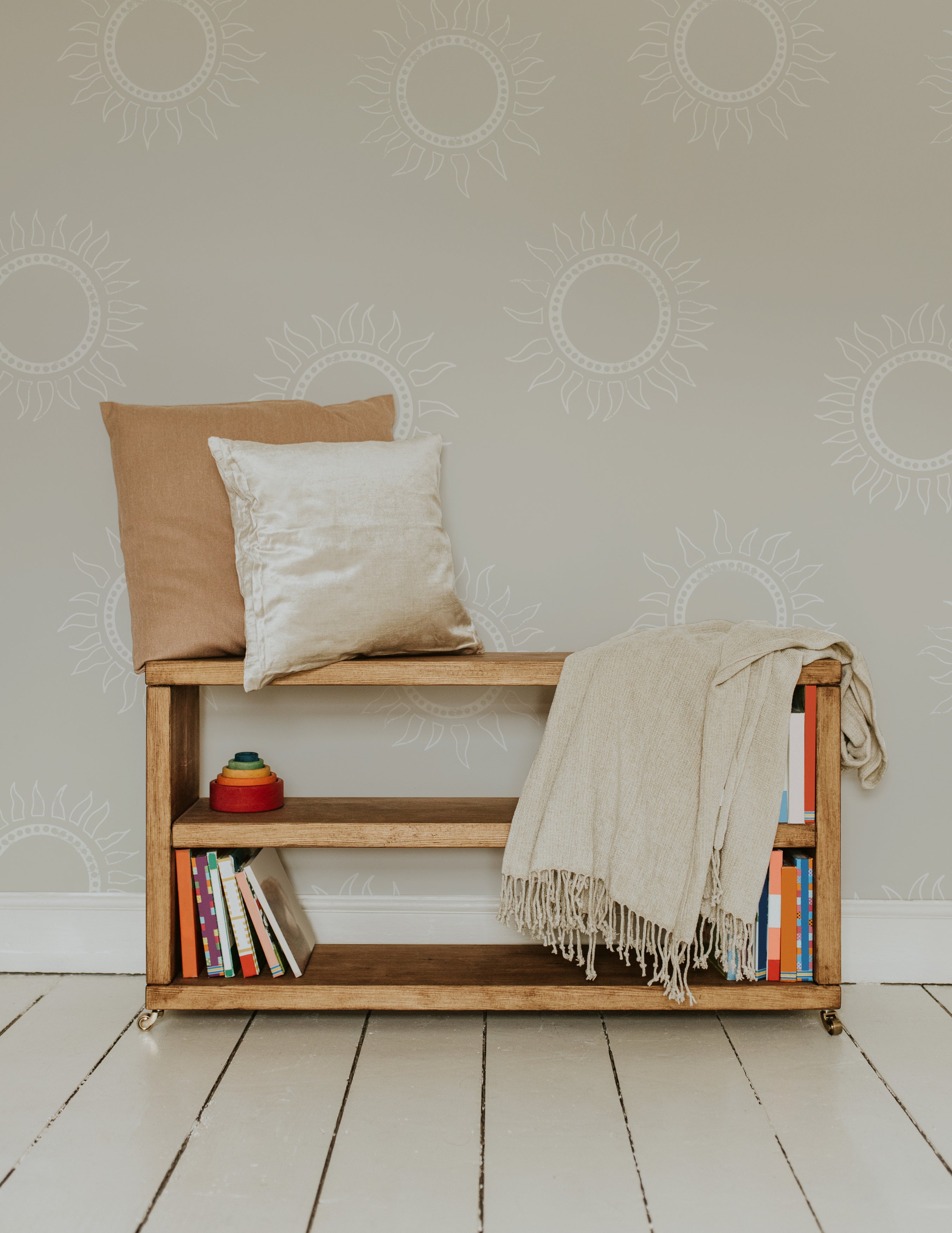  A cozy corner with the Sunburst Harmony Wallpaper on the wall, featuring the white sunburst pattern on a warm beige background. A wooden bench with pillows and a blanket is placed against the wall, with books neatly arranged on the lower shelves.