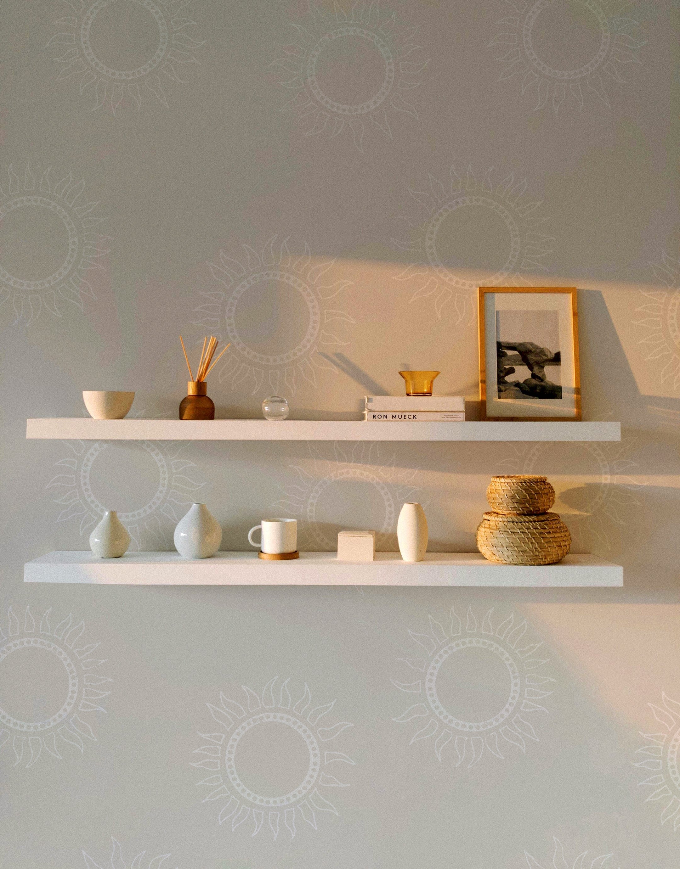 A room featuring Sunburst Harmony Wallpaper in a warm beige color. The wallpaper displays a subtle sunburst pattern in white. Two floating shelves hold various decorative items, including vases, a framed picture, and a small bowl, creating a minimalist and cozy look.