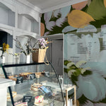 A cozy café corner where the Summer Floral Mural Wallpaper creates a vivid statement wall with its oversized, abstract flower illustrations in shades of yellow, blue, and white against a dark green backdrop, adding a dramatic yet playful atmosphere to the dessert showcase area.
