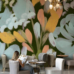 A woman sits comfortably in a café with the Summer Floral Mural Wallpaper adorning the wall, the large, abstract floral design enveloping the space in a cascade of cool and warm tones, offering a vibrant and artistic backdrop for a relaxing ambiance.