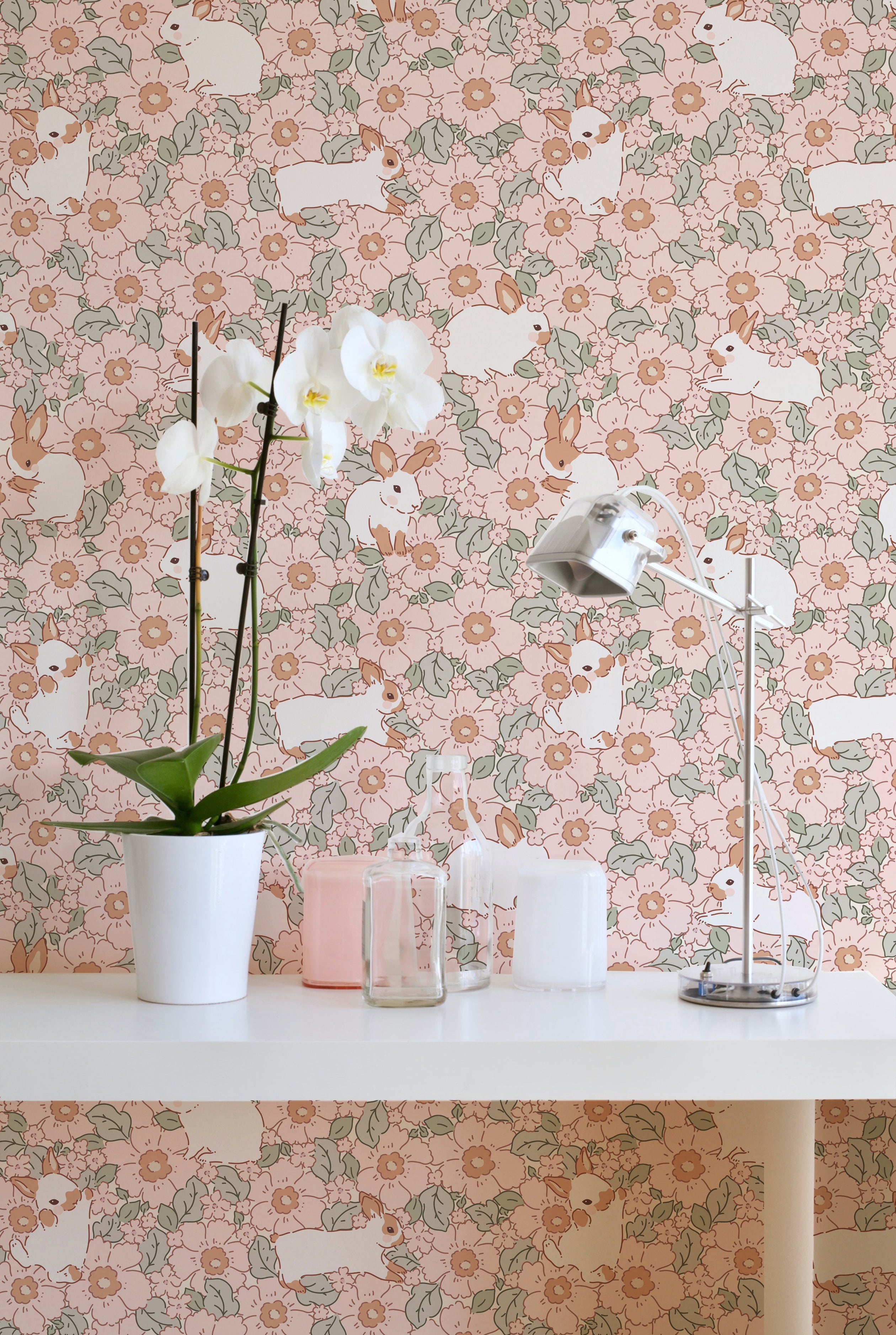 A bright workspace is enhanced by the Petite Lapinou Wallpaper, displaying a delightful pattern of white rabbits and floral motifs in soft pink and green. The desk is adorned with a white orchid, modern lamp, and minimalist accessories, creating a peaceful and stylish area for creativity.