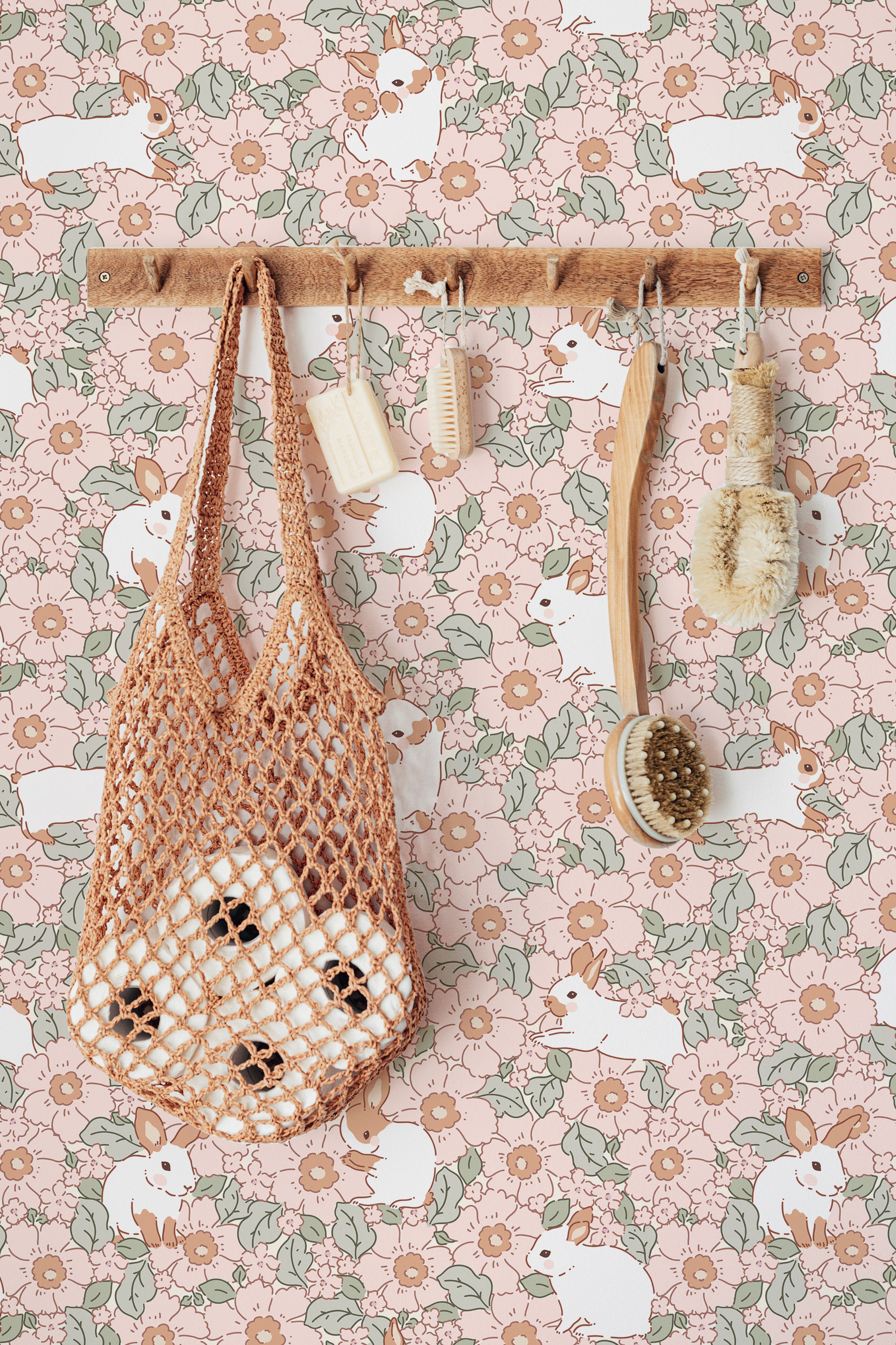 A charming scene showcasing the Petite Lapinou Wallpaper, adorned with whimsical illustrations of white rabbits amidst a floral pattern of pink and green. The wallpaper serves as a backdrop for a wooden rack hung with a netted bag and wooden brushes, enhancing the pastoral and serene feel of the setting.