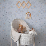 A nursery room enhanced with the Blue Floral Joy Wallpaper, featuring small white floral motifs and playful celestial symbols on a light blue backdrop. The room includes a wicker bassinet filled with plush toys, creating a warm and inviting space for a child
