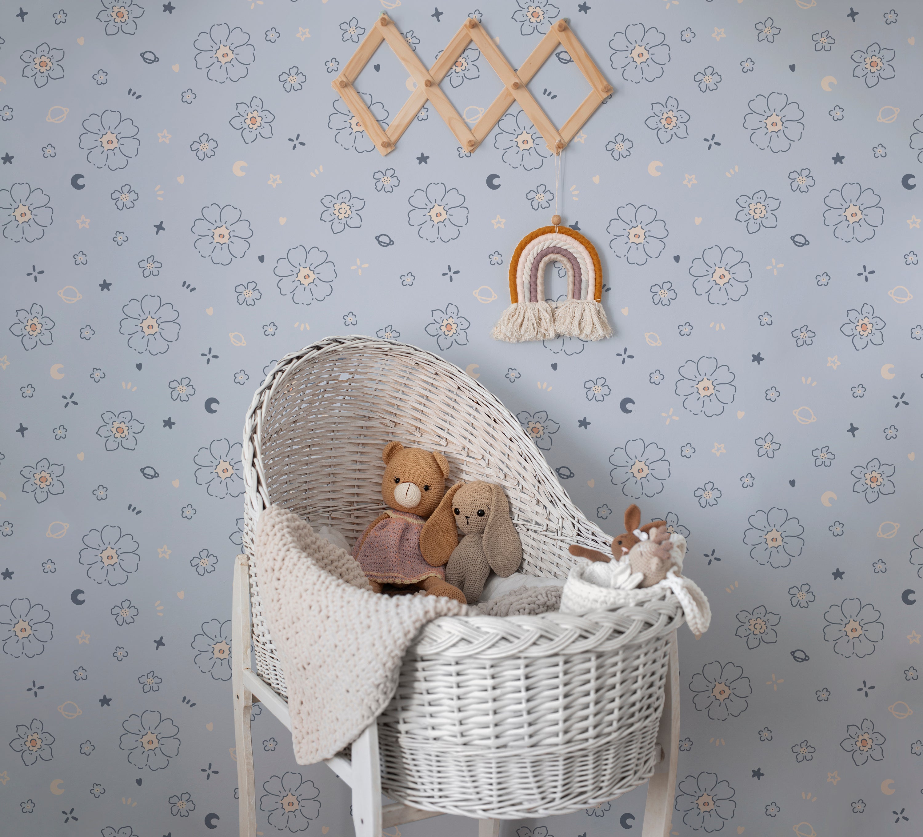 A nursery room enhanced with the Blue Floral Joy Wallpaper, featuring small white floral motifs and playful celestial symbols on a light blue backdrop. The room includes a wicker bassinet filled with plush toys, creating a warm and inviting space for a child
