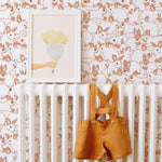 A cozy nursery scene featuring Lapinou Wallpaper with a pattern of playful beige bunnies on a cream background. A white crib is against the wall, adorned with a framed art piece of a hand holding a bouquet and a pair of mustard yellow toddler suspenders hanging on the crib rail.