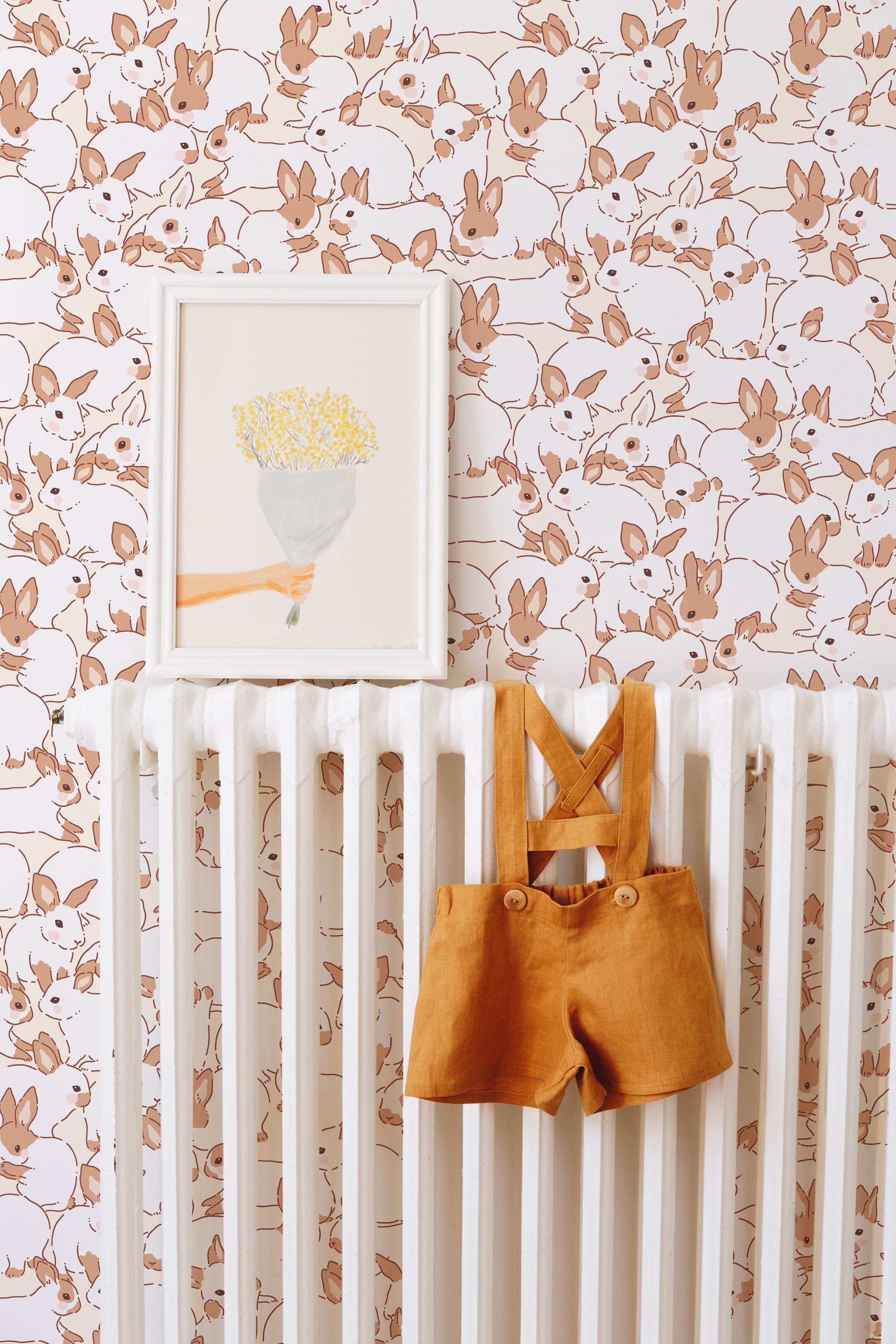 A cozy nursery scene featuring Lapinou Wallpaper with a pattern of playful beige bunnies on a cream background. A white crib is against the wall, adorned with a framed art piece of a hand holding a bouquet and a pair of mustard yellow toddler suspenders hanging on the crib rail.