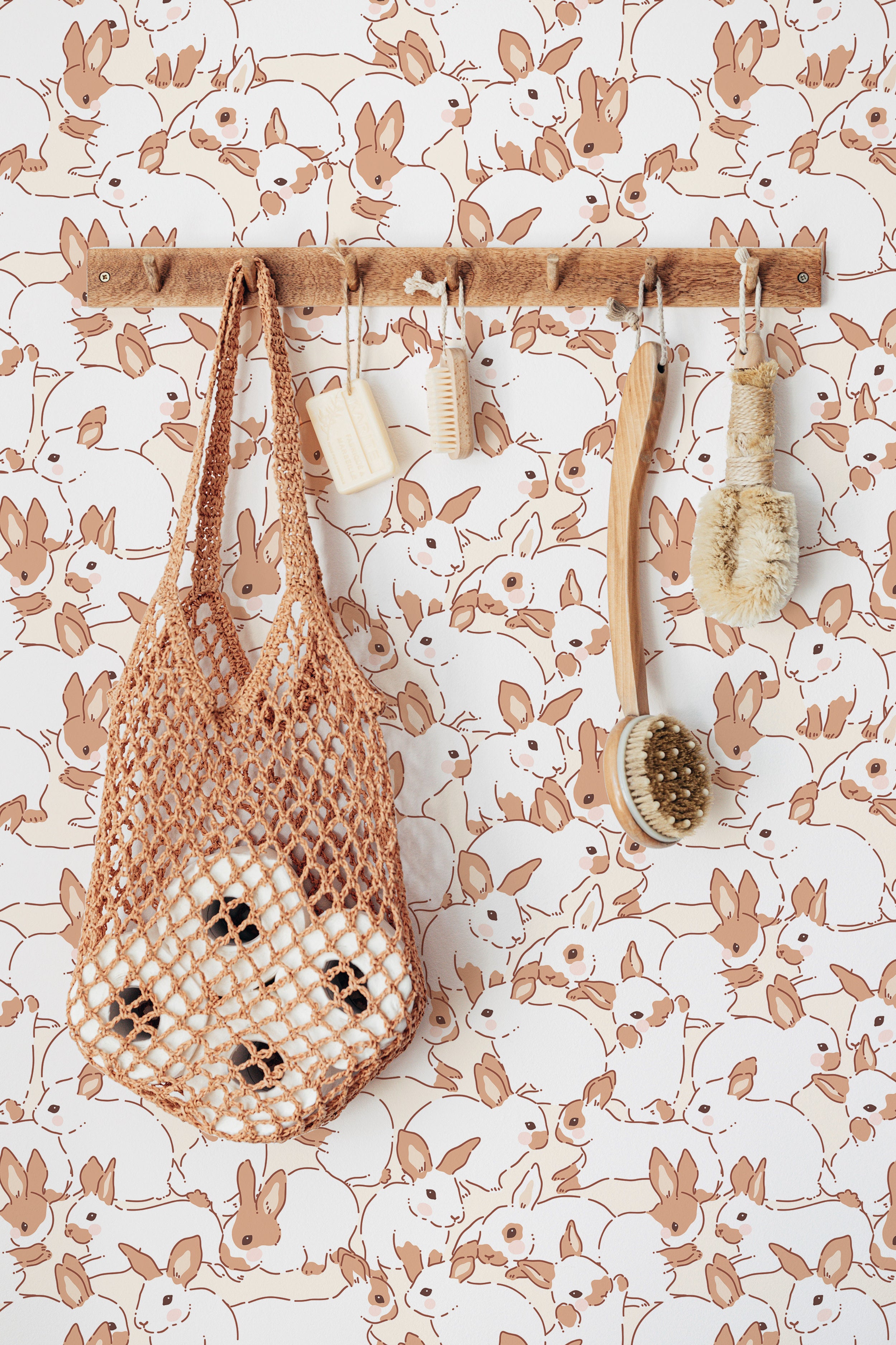 Interior decor scene showcasing the Lapinou Wallpaper with beige bunny patterns. A wooden rack holds various natural bath accessories including a mesh bag, a wooden brush, and two bars of soap, enhancing the rustic charm of the room.
