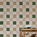 A room featuring Clémence Wallpaper II with a checkered pattern in shades of green, beige, pink, and white. The wallpaper covers the wall behind a wooden shelf with books and toys, including a white toy truck.