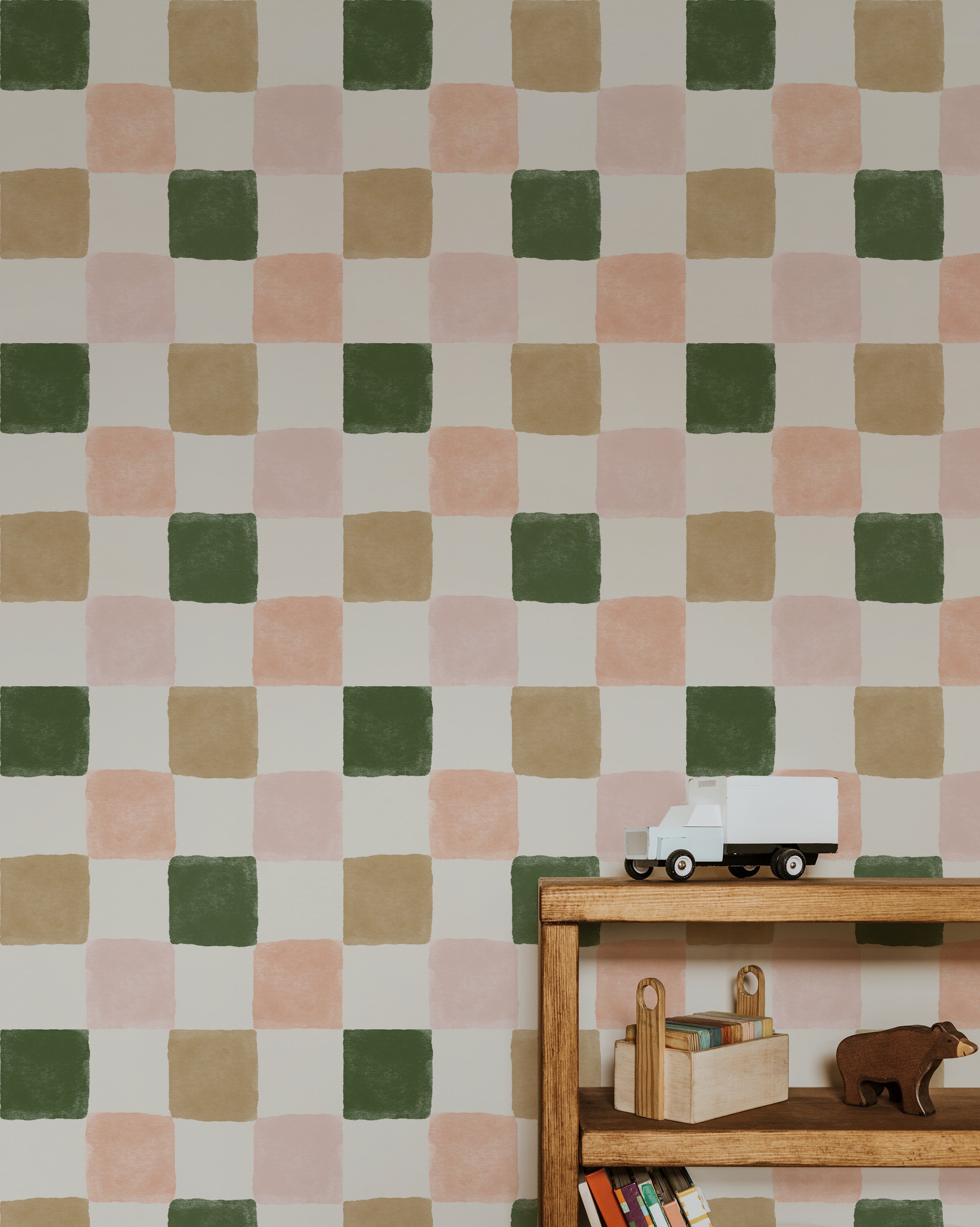 A room featuring Clémence Wallpaper II with a checkered pattern in shades of green, beige, pink, and white. The wallpaper covers the wall behind a wooden shelf with books and toys, including a white toy truck.