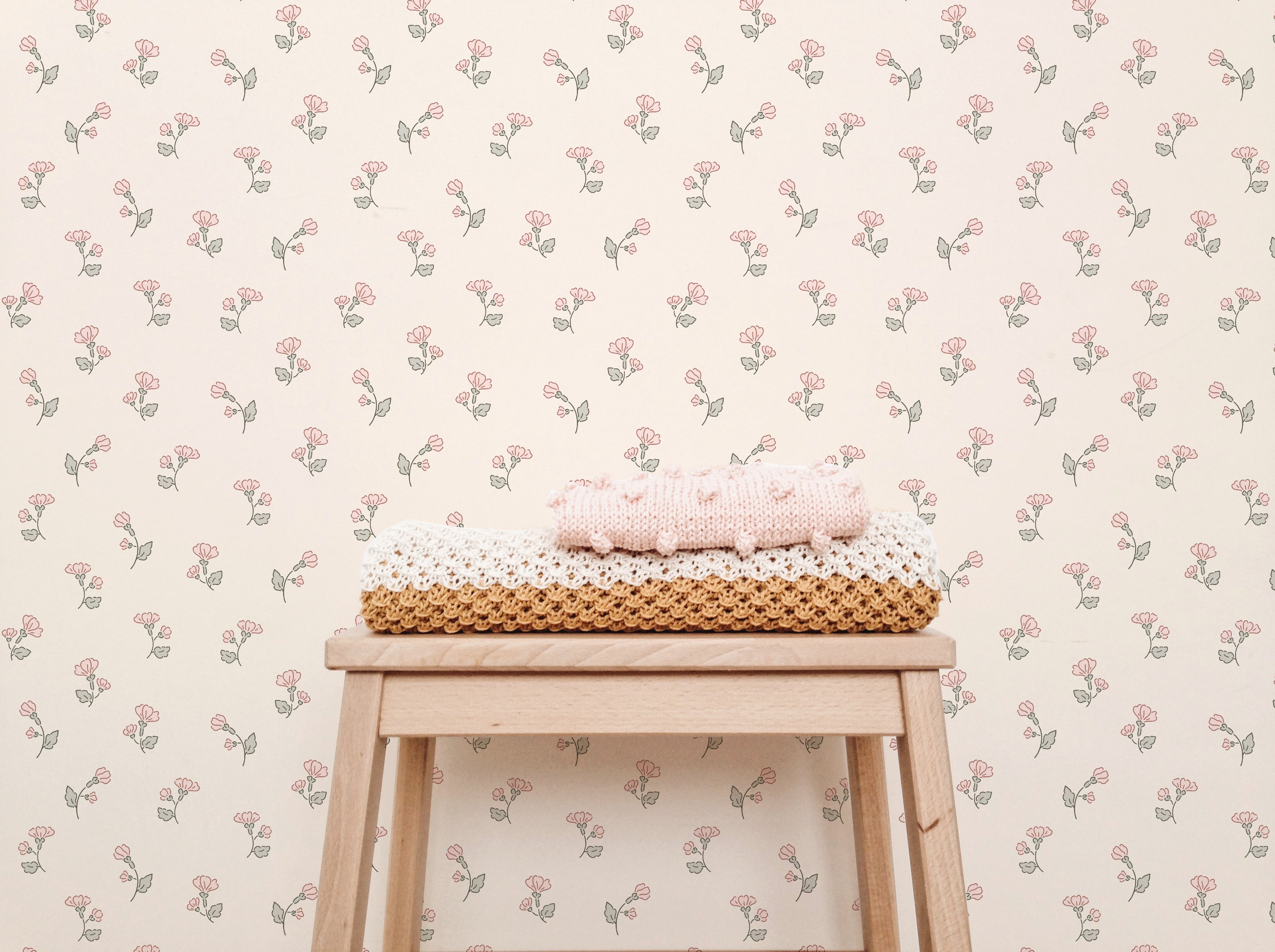 A gentle and inviting nursery scene with Chara Wallpaper, which displays a delicate pattern of small pink and green floral sprigs on a soft peach background. A wooden stool is placed against the wall, topped with a stack of hand-knitted blankets in pink, white, and brown, adding a cozy touch to the decor.