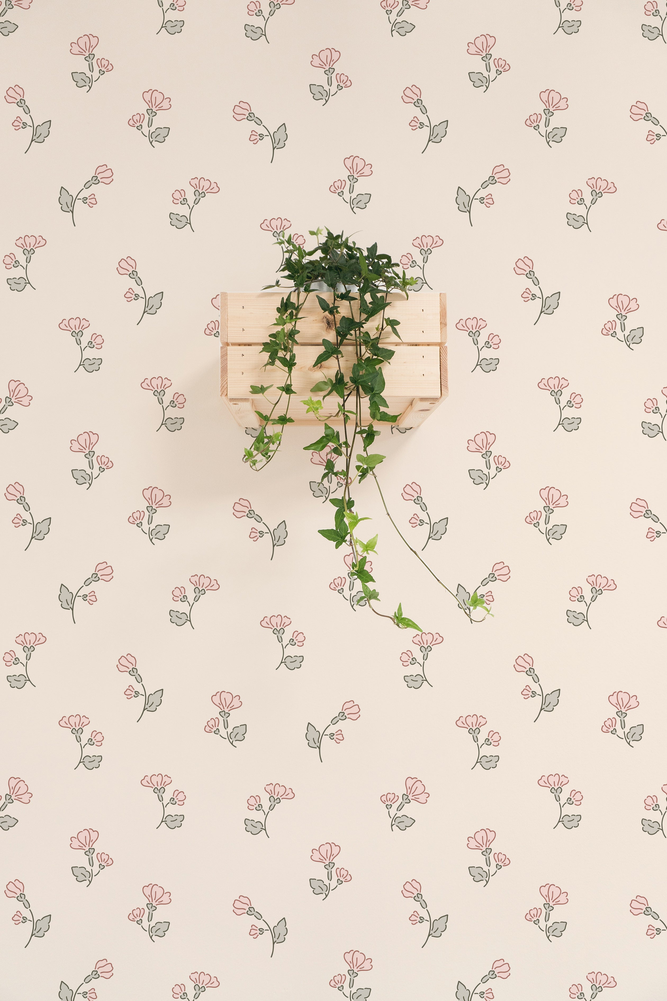 An elegant interior scene featuring Chara Wallpaper with a charming pattern of pink floral blooms and subtle green foliage on a cream background. A wooden box shelf on the wall is adorned with a lush green plant draping over the sides, complementing the floral theme of the wallpaper.