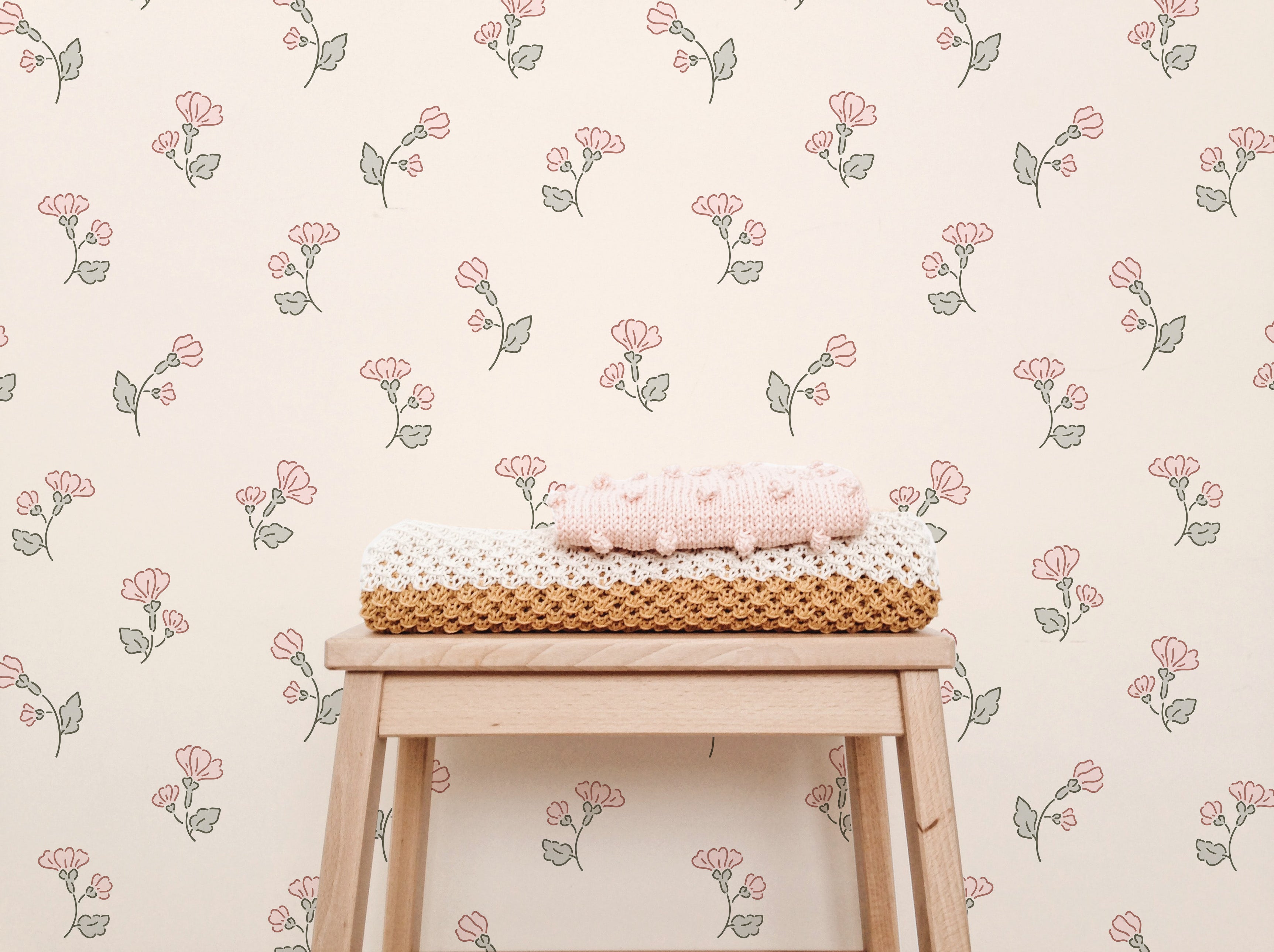 A cozy nursery setting enhanced by the Chara Wallpaper, which showcases delicate pink flowers and green leaves on a cream backdrop. A wooden stool displays a stack of hand-knitted blankets in shades of pink, white, and brown, adding a touch of homely warmth against the floral wallpaper.