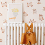 A charming nursery setting showcasing the Chaton Wallpaper, featuring adorable beige and brown kittens on a light peach background. The room includes a white crib, above which hangs a framed artwork of a yellow bouquet, complemented by mustard-colored toddler suspenders draped over the crib rail.