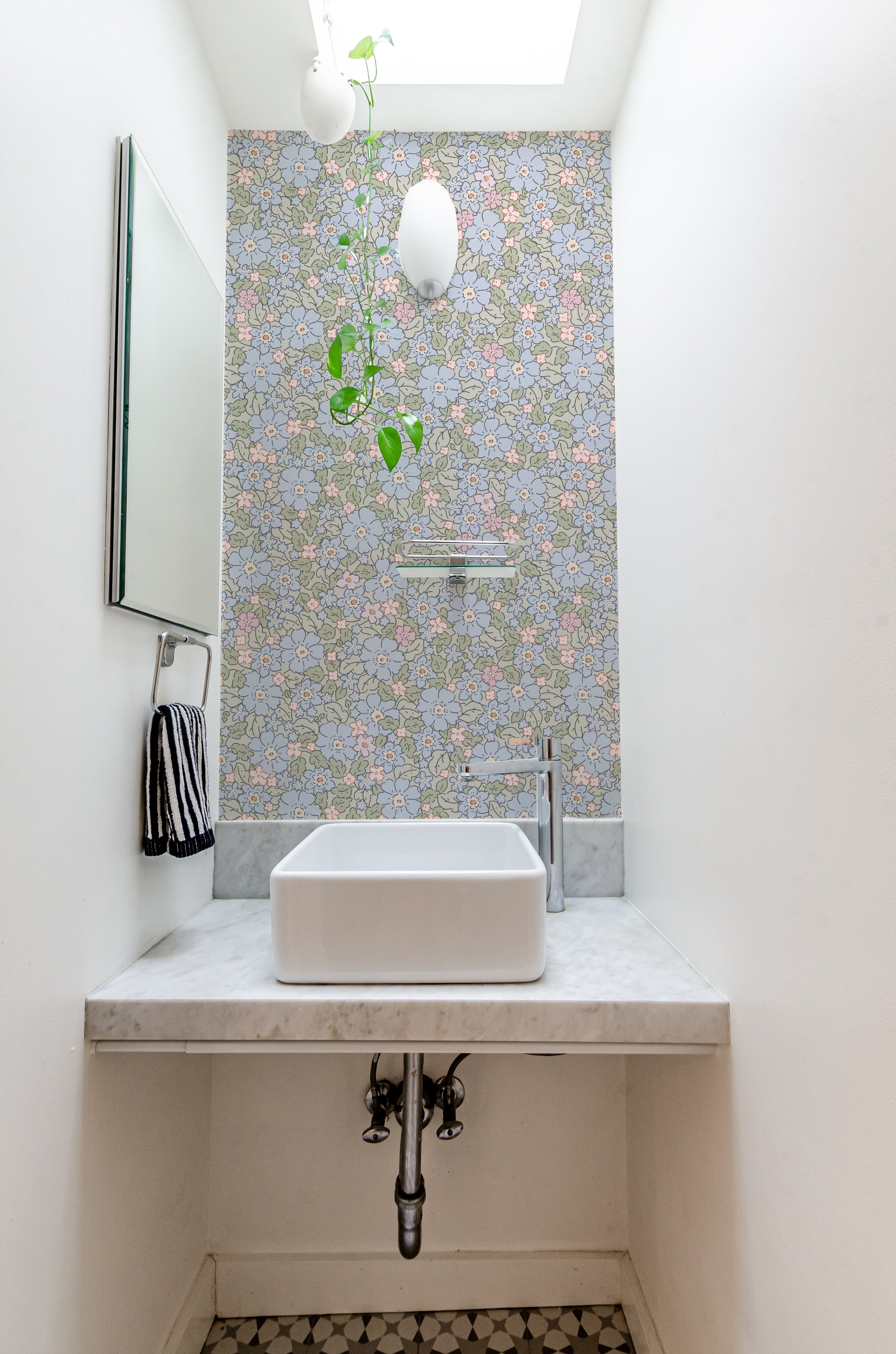 A small and elegant bathroom space enhanced with Joie Wallpaper, which features a beautiful floral pattern in shades of blue, pink, and green on a light green background. The room is fitted with a modern white sink on a marble countertop, under a skylight that adds natural light to the floral ambiance.