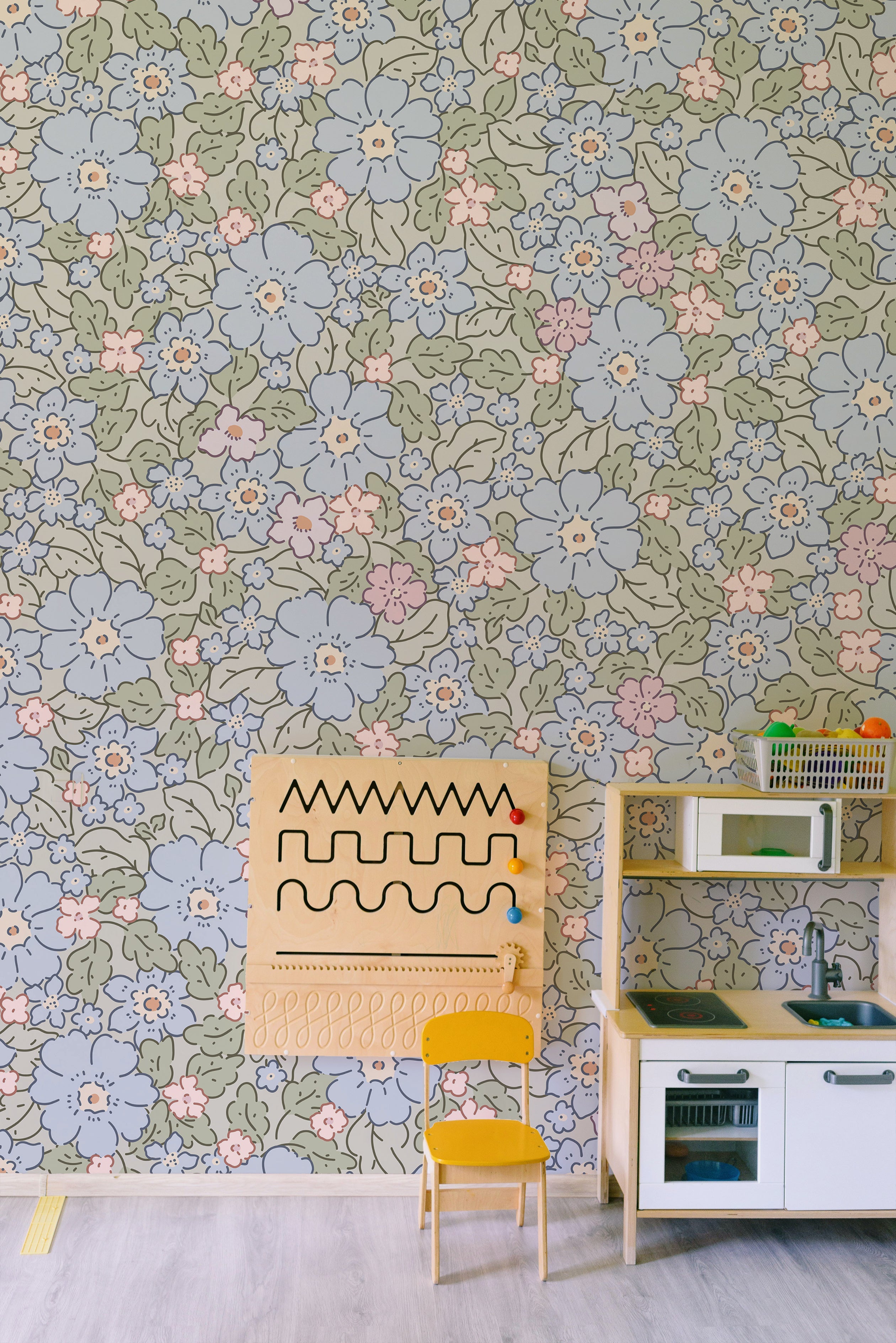 A playful and colorful children's play area decorated with Joie Wallpaper, which displays a cheerful floral pattern in soft blue, pink, and green. The room includes a large wooden activity board, a bright yellow chair, and a child-sized kitchen playset, providing a stimulating and inviting space for children.