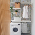 A modern laundry area featuring Joie Wallpaper with a vibrant floral pattern of blue, pink, and green flowers on a light green background. The space is efficiently organized with a white washing machine, built-in shelving holding various baskets and green plants, and white shirts hanging on wooden hangers, creating a refreshing and functional environment.