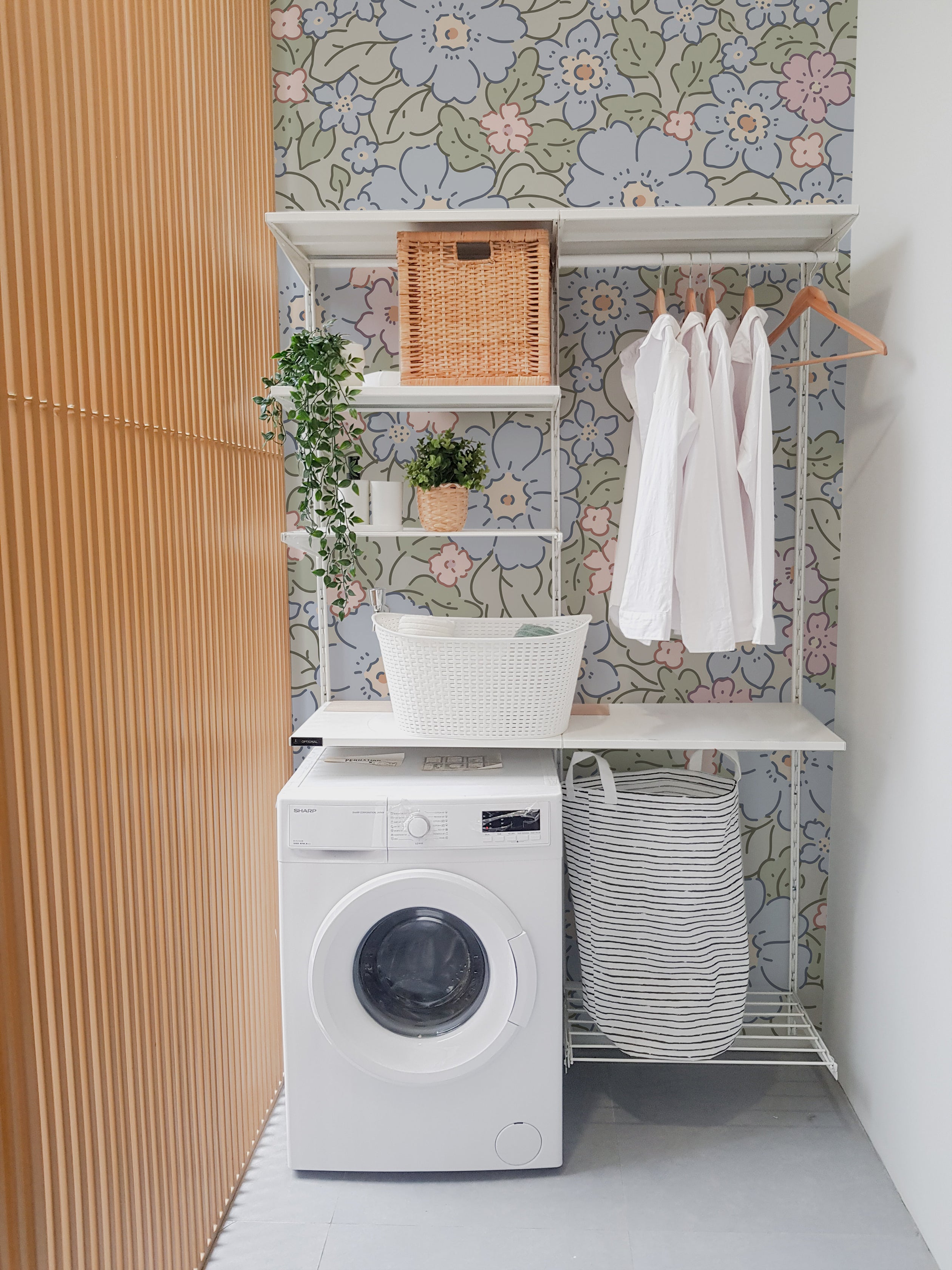 A modern laundry area featuring Joie Wallpaper with a vibrant floral pattern of blue, pink, and green flowers on a light green background. The space is efficiently organized with a white washing machine, built-in shelving holding various baskets and green plants, and white shirts hanging on wooden hangers, creating a refreshing and functional environment.