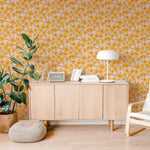 A contemporary home office setup featuring a light wooden sideboard against a vibrant Abstract Meadow Wallpaper. The wallpaper displays a pattern of small yellow and peach flowers on a pale pink background, creating a fresh and cheerful ambiance. The scene is completed with a lush potted plant, a stylish white lamp, and cozy seating, enhancing the room’s natural light and spacious feel.