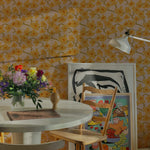 A cozy dining area adorned with Abstract Meadow Wallpaper that brightens the space with its pattern of yellow flowers on a pink background. The room features a circular wooden table, eclectic chairs, and a vibrant mix of art prints and flowers, creating a welcoming and artistic dining experience.