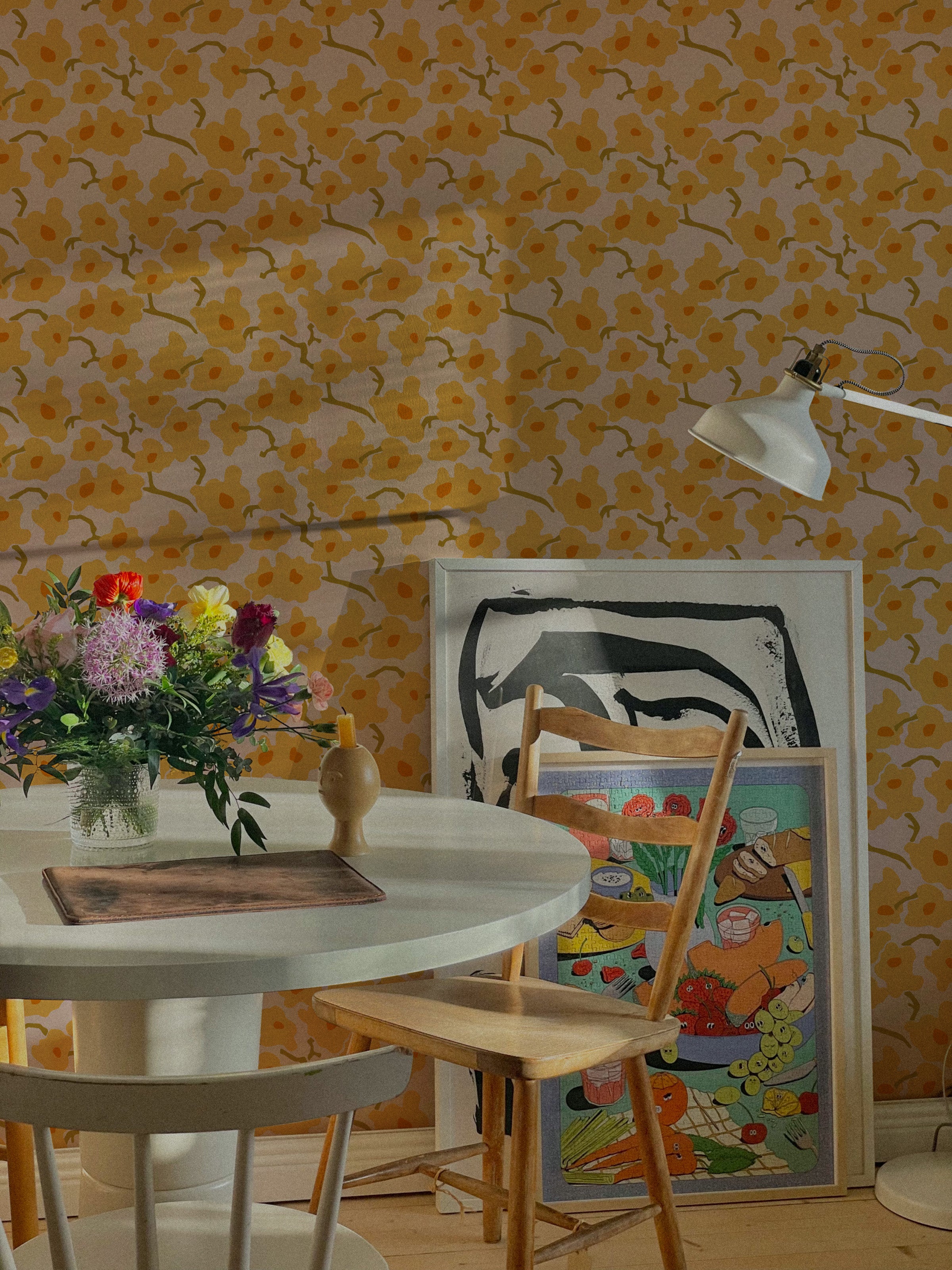 A cozy dining area adorned with Abstract Meadow Wallpaper that brightens the space with its pattern of yellow flowers on a pink background. The room features a circular wooden table, eclectic chairs, and a vibrant mix of art prints and flowers, creating a welcoming and artistic dining experience.
