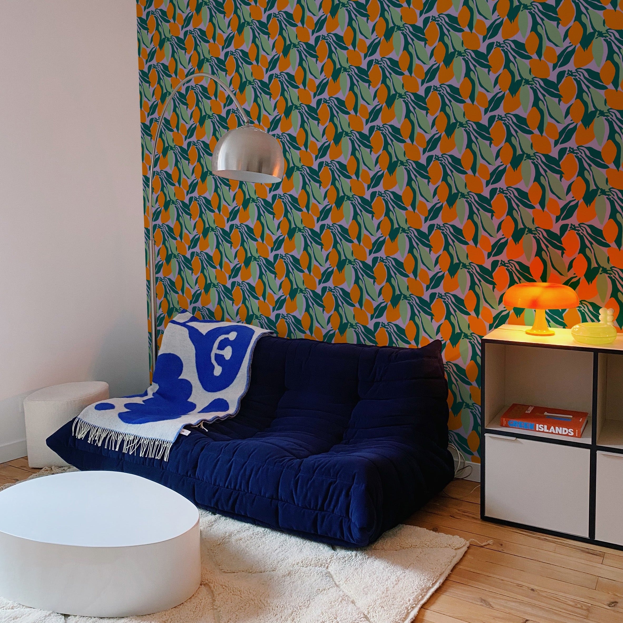 A modern living room featuring a wall covered in wallpaper with a pattern of orange fruits and green leaves on a purple background. The room includes a navy blue sofa, a round white coffee table, and various modern decorations, creating a vibrant and stylish space