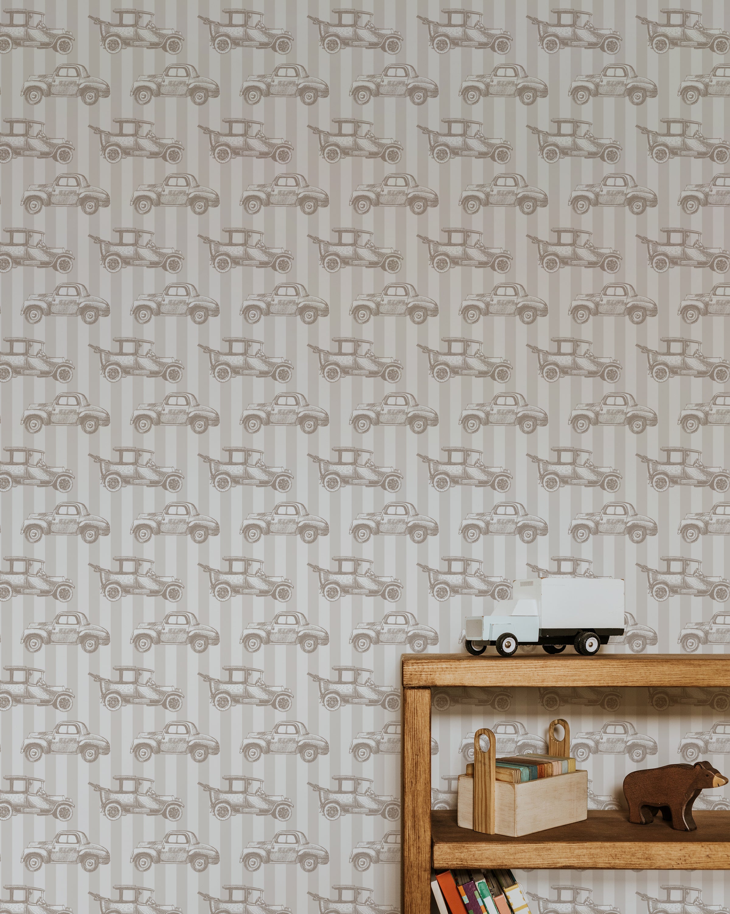 A full view of a wall covered with Antique Auto Wallpaper in beige. The design features intricate illustrations of classic cars, creating a nostalgic and vintage ambiance. A wooden shelf with toys and books is positioned against the wall.