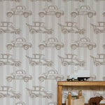 A wooden bookshelf with books and toys set against a wall covered in Antique Auto Wallpaper. The wallpaper features a whimsical pattern of vintage cars on a beige striped background