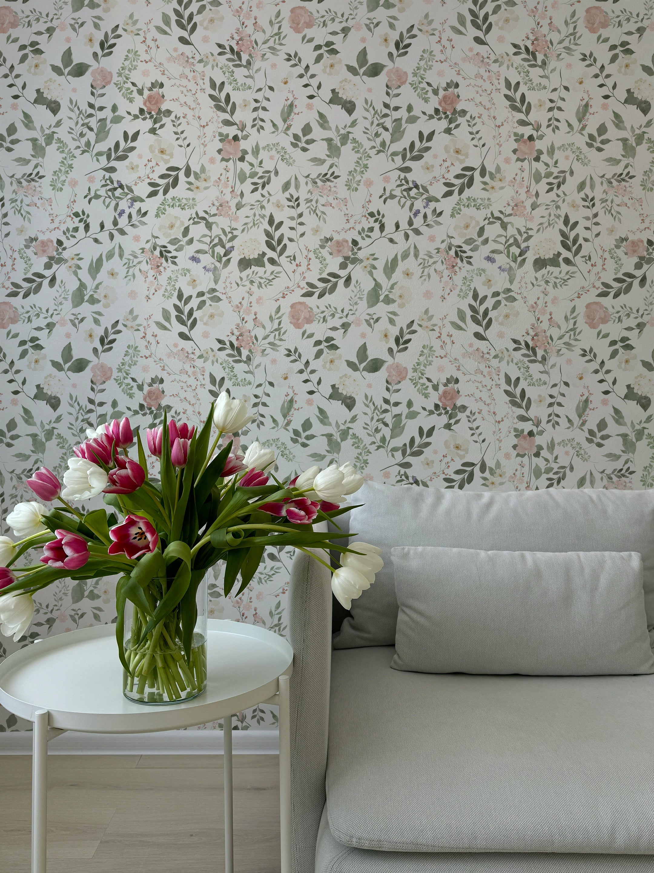 A modern living room with a light gray sofa and a vase of tulips on a side table, set against a wall decorated with Enchanted Symphony Wallpaper. The wallpaper displays a subtle and graceful floral design with green foliage and pink blooms.